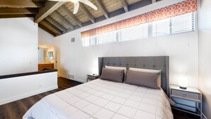 Un dormitorio con una cama grande, vigas de madera en el techo, una mesita de noche con lámpara y una ventana con persianas.