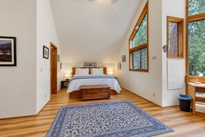 A bedroom with a large bed, wooden flooring, a blue patterned rug, and a window with blinds. - Spacious dog-friendly cabin with creek & mountain views (Idyllwild)