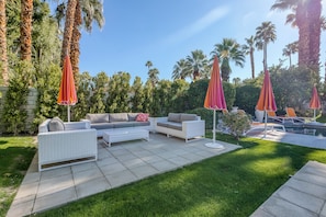 A patio area with outdoor furniture and umbrellas, surrounded by palm trees and a pool. - Beautiful home w/ mountain views, private pool & gas fireplace (Palm Springs)