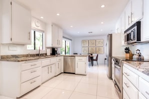 A modern kitchen with white cabinetry, a dishwasher, a microwave, and a coffee maker. There is a dining area with a table and chairs visible in the background. - Beautiful home w/ mountain views, private pool & gas fireplace (Palm Springs)