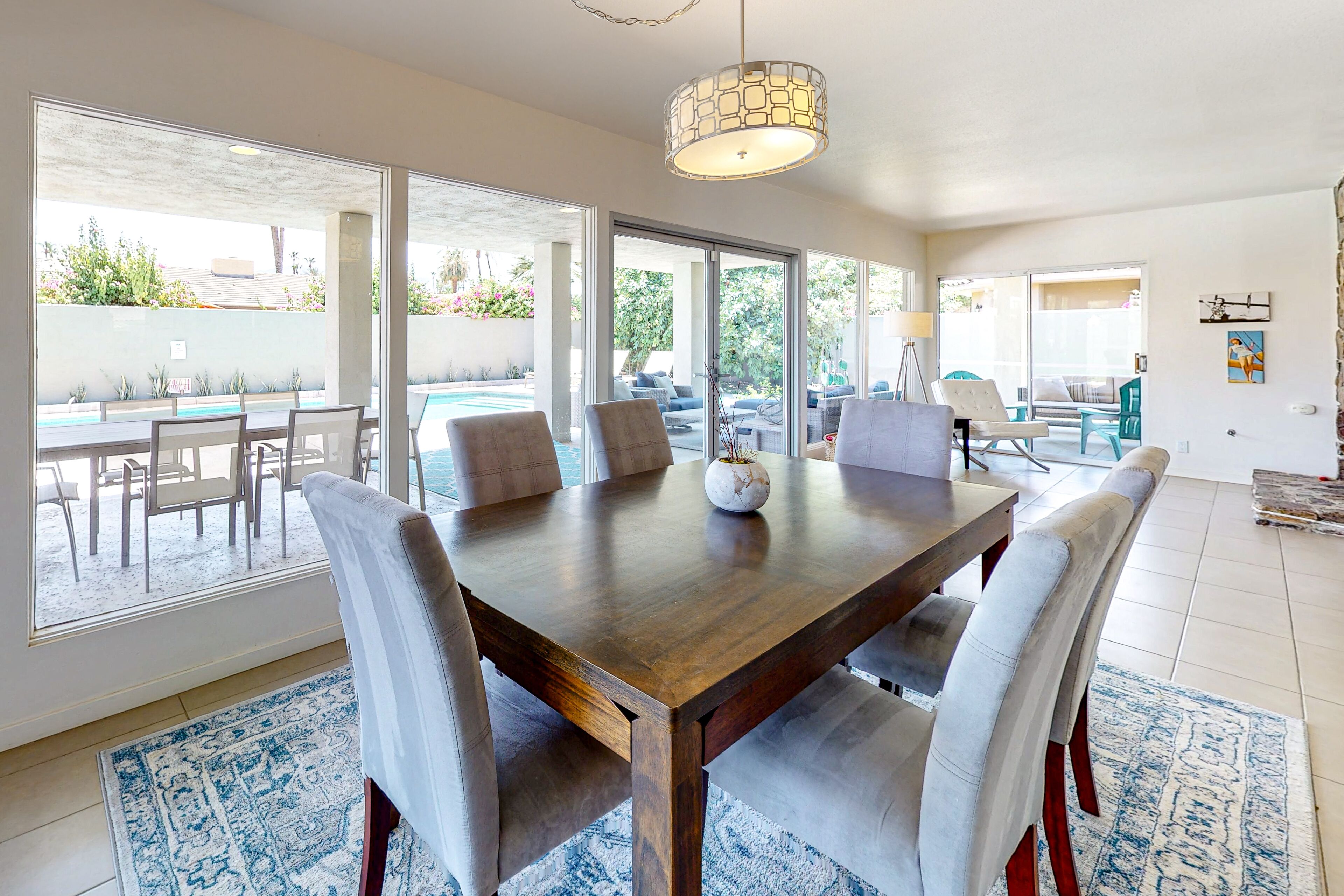 A spacious dining area with a large wooden table and six chairs, large windows offering a view of the outdoors, and a hanging light fixture.