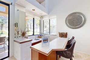 A modern kitchen with a large island, a dining area with wicker chairs, and a decorative metal plate on the wall. - Lavish condo on 14th tee of Arnold Palmer course at PGA West (La Quinta)
