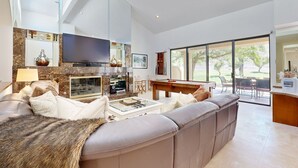 Uma sala de estar moderna com um amplo sofá seccional, uma TV de tela plana, lareira e uma área de jantar com vista para o exterior.