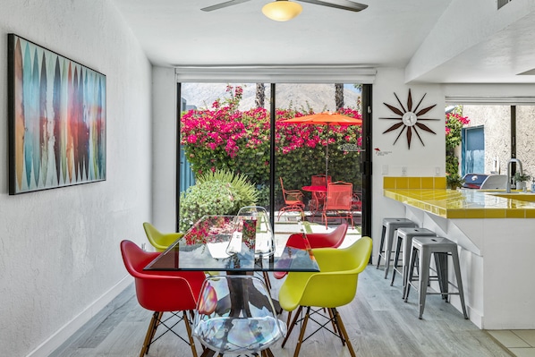 Una cocina moderna con una mesa de vidrio y sillas coloridas, un salpicadero amarillo y vistas a un jardín con flores rosadas.