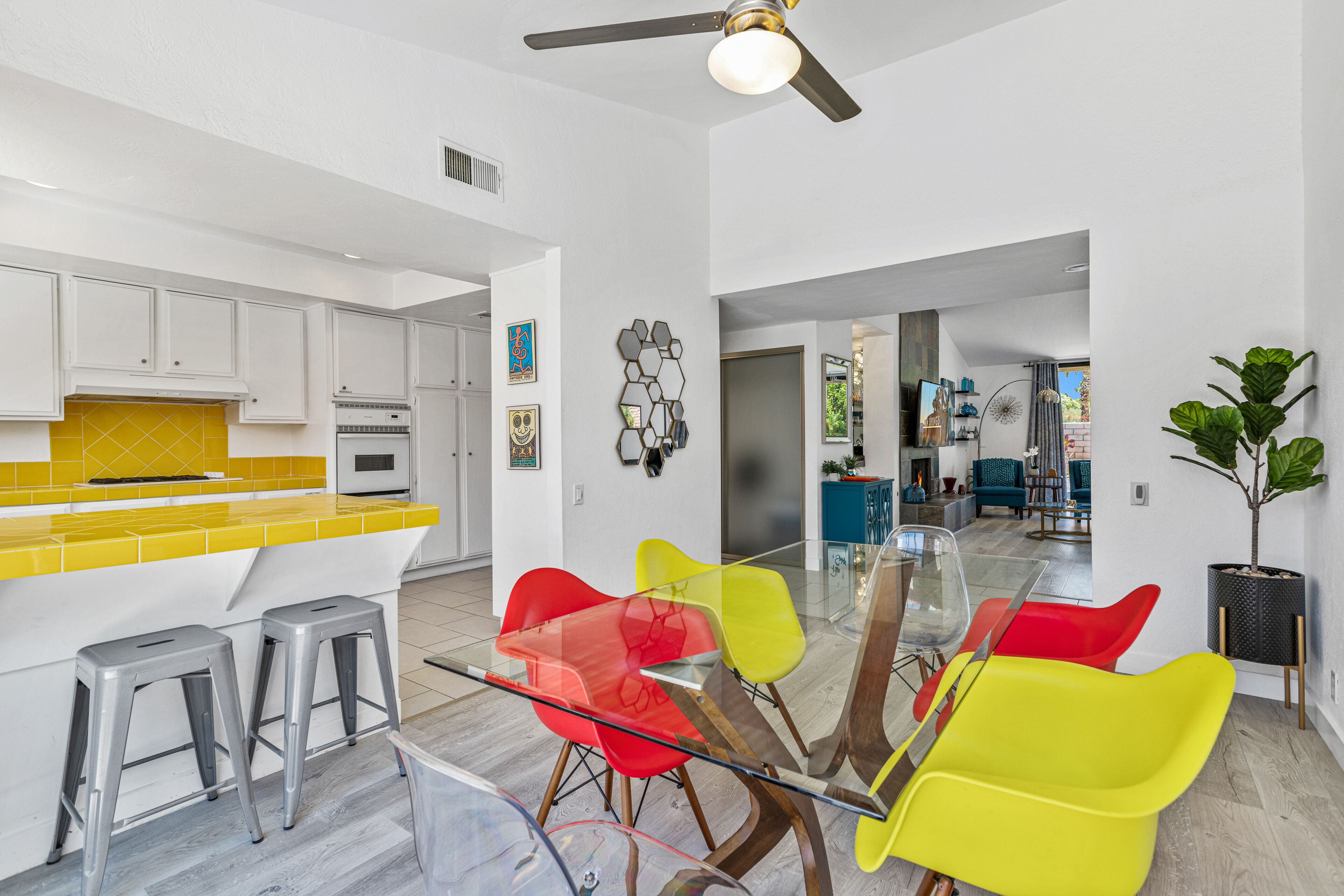 A modern kitchen with white cabinets, a yellow backsplash, and a dining area with a glass table and colorful chairs.