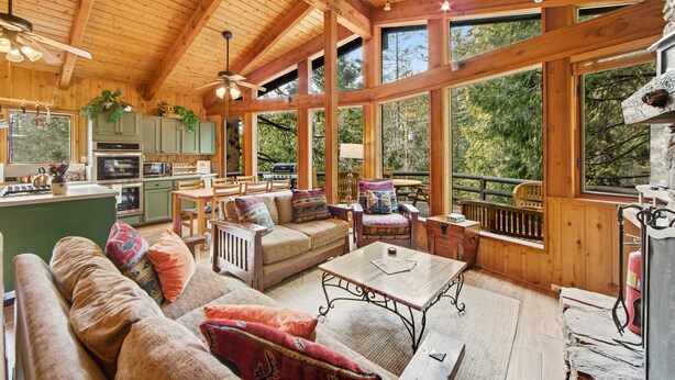 A spacious living area with a wooden ceiling, large windows, a kitchen, and a cozy seating arrangement.