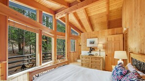 A wooden cabin bedroom with a bed, dresser, and large windows overlooking a forest.