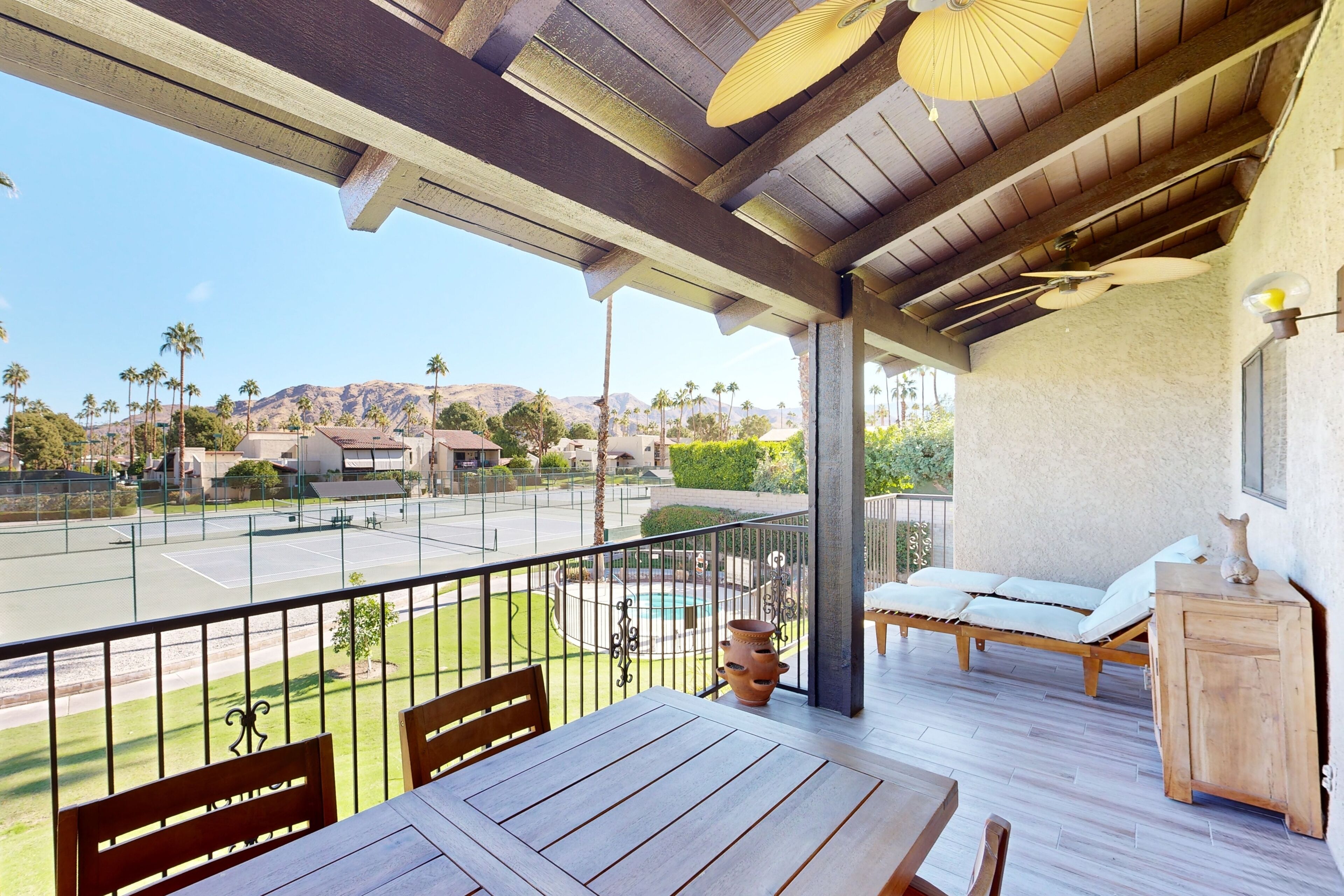 A balcony with a wooden table and chairs, a lounge chair, and a view of a tennis court and pool area.
