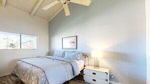 Un dormitorio con cama, mesita de noche, lámpara, ventilador de techo y ventana con persianas.