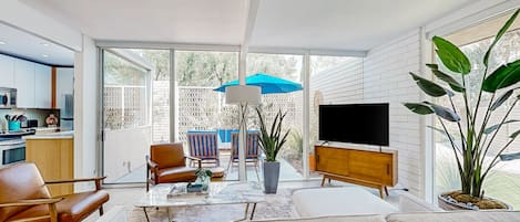 Sala de estar moderna com sofá, mesa de centro e televisão. Há uma área de cozinha com armários e micro-ondas. Lá fora, existe um pátio com cadeiras e um guarda-sol azul.