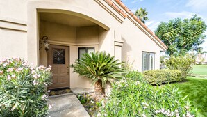 Een huis met een beige gevel, een bruine deur en een kleine tuin met diverse planten en bloemen.