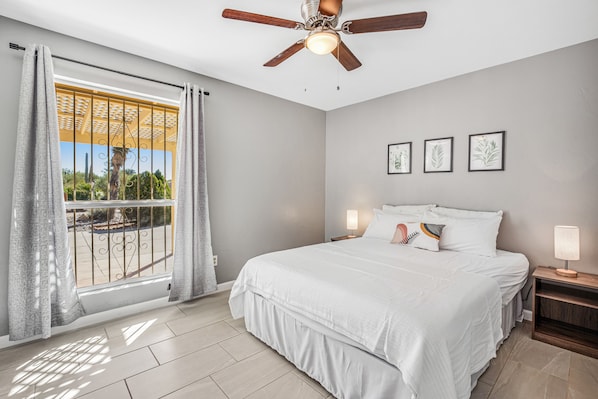A bedroom with a bed, a ceiling fan, a nightstand with a lamp, and a window with curtains. - Dog-friendly sunshine stay with private pool, spacious yard, central AC & W/D (Tucson)