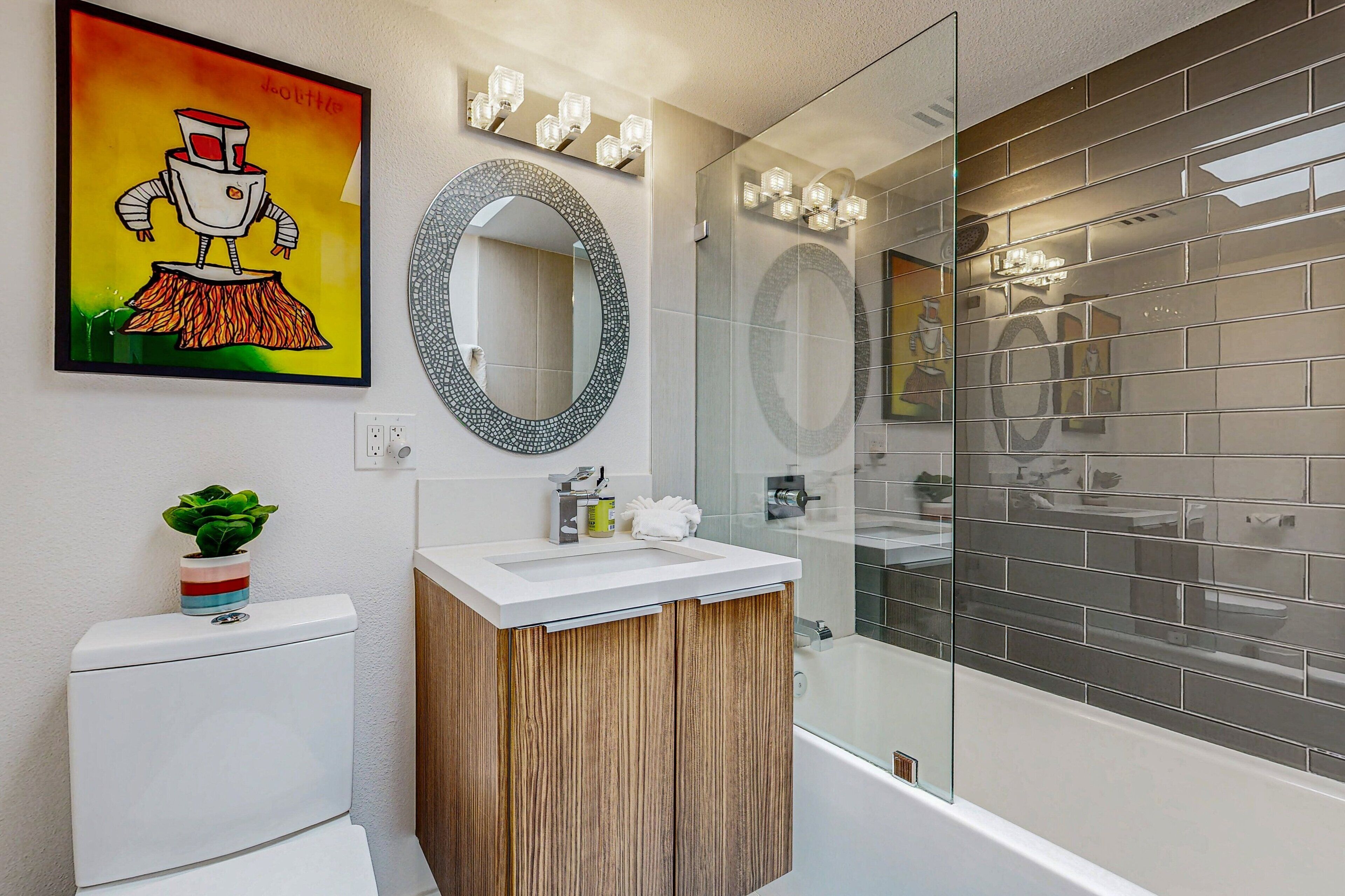 A modern bathroom with a toilet, sink, and bathtub. There is a mirror above the sink and a large mirror above the bathtub. A colorful painting is on the wall above the toilet.