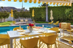 Zona de piscina al aire libre con mesa de comedor para cuatro personas, rodeada de sillas amarillas y un área de descanso con sillas y sombrillas.