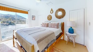 A bedroom with a bed, a ceiling fan, a view of the outdoors, a door, and a small table with a lamp. - Secluded hilltop home with hot tub, cowboy pool, & amazing views (Yucca Valley)