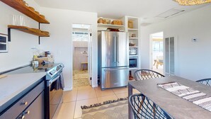 A modern kitchen with a stainless steel refrigerator, wooden shelves, and a dining area with a table and chairs. - Secluded hilltop home with hot tub, cowboy pool, & amazing views (Yucca Valley)
