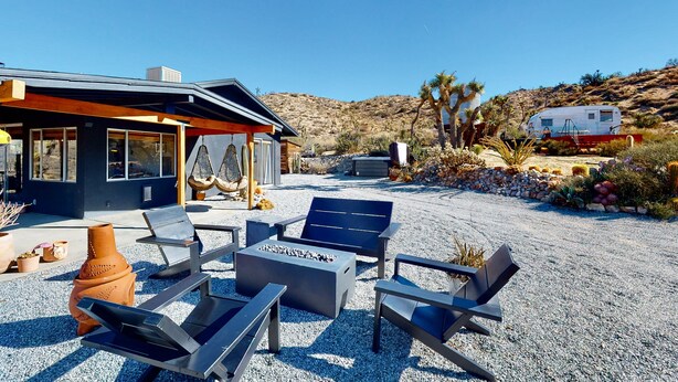 A modern outdoor seating area with a fire pit, surrounded by a gravel patio, and a desert landscape in the background. - Secluded hilltop home with hot tub, cowboy pool, & amazing views (Yucca Valley)