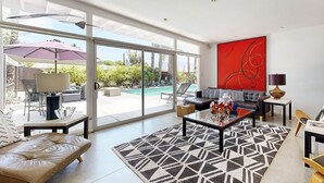 A spacious living room with a large glass door leading to a pool area, a modern sofa, a coffee table, and a vibrant abstract painting on the wall. - Mid-Century Jack LaLanne home with pool, spa & firepit (Palm Springs)