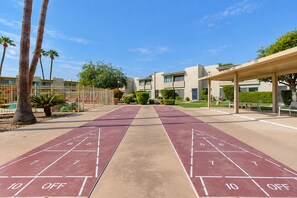 A shuffleboard court with a clear sky, palm trees, and a building with a covered seating area. - Welcoming condo with WiFi & pool - near Scottsdale Fashion Square (Scottsdale)