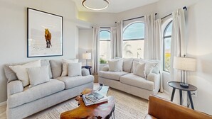 A modern living room with two sofas, a coffee table, and a lamp. There are large windows with curtains, a framed picture on the wall, and a ceiling light fixture. - Luxury home w/ private pool & optional pool heat, rooftop patio & mountain views (Fountain Hills)