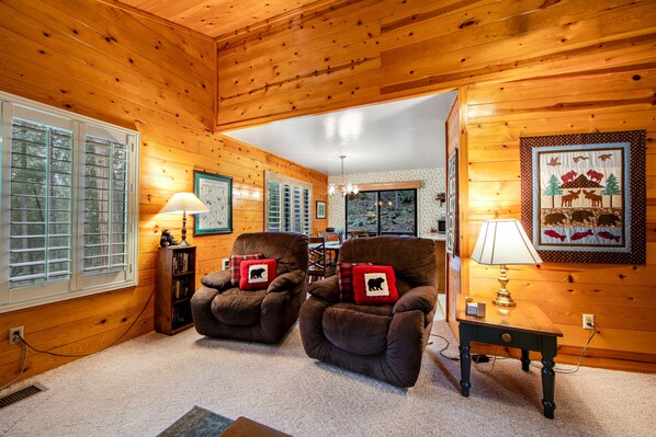 A cozy cabin interior with wooden walls, two armchairs with bear-themed pillows, a table with a lamp, and a framed wildlife scene on the wall. - Secluded Dog-Friendly Cabin w/Wood Fireplace, Deck, Private W/D, Mtn Views (Idyllwild)