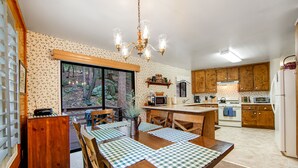 A kitchen with wooden cabinets, a dining table with checkered placemats, and a large window overlooking a forest. - Secluded Dog-Friendly Cabin w/Wood Fireplace, Deck, Private W/D, Mtn Views (Idyllwild)