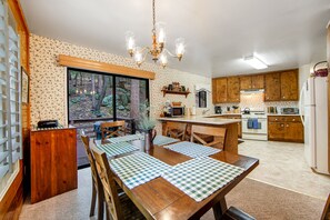 A kitchen with wooden cabinets, a dining table with checkered placemats, and a large window overlooking a forest. - Secluded Dog-Friendly Cabin w/Wood Fireplace, Deck, Private W/D, Mtn Views (Idyllwild)