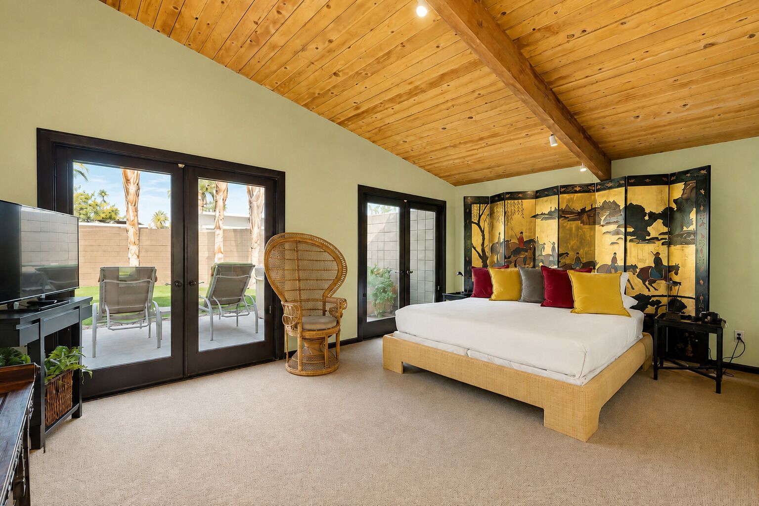 Een slaapkamer met een groot bed, een televisie, een rieten fauteuil en een glazen schuifdeur die leidt naar een buitenruimte.