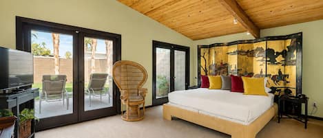 A bedroom with a large bed, a television, a rattan chair, and a sliding glass door leading to an outdoor area.