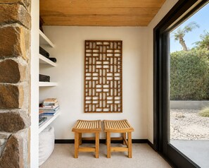 Une pièce avec un mur en pierre, un plafond en bois, une bibliothèque, deux tabourets en bois et une vue sur un palmier par la fenêtre.