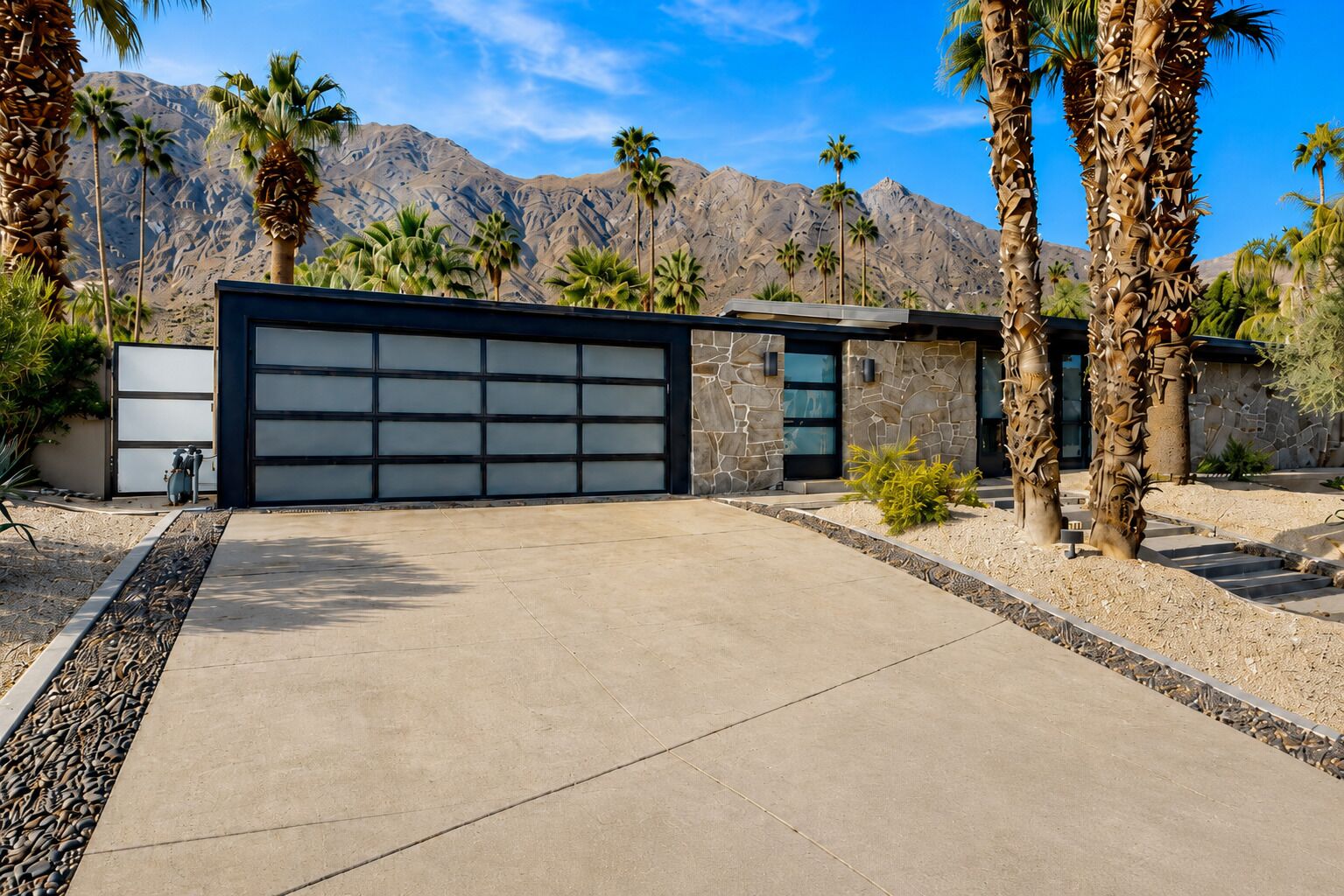 A modern house with a garage, palm trees, and mountains in the background.