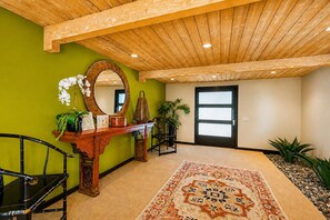 A room with a wooden ceiling, a mirror, a door, a console table with decorative items, a patterned rug, and a chair. - Stylish Mid-century gem near downtown with pool, hot tub, fire table & WiFi (Palm Springs)