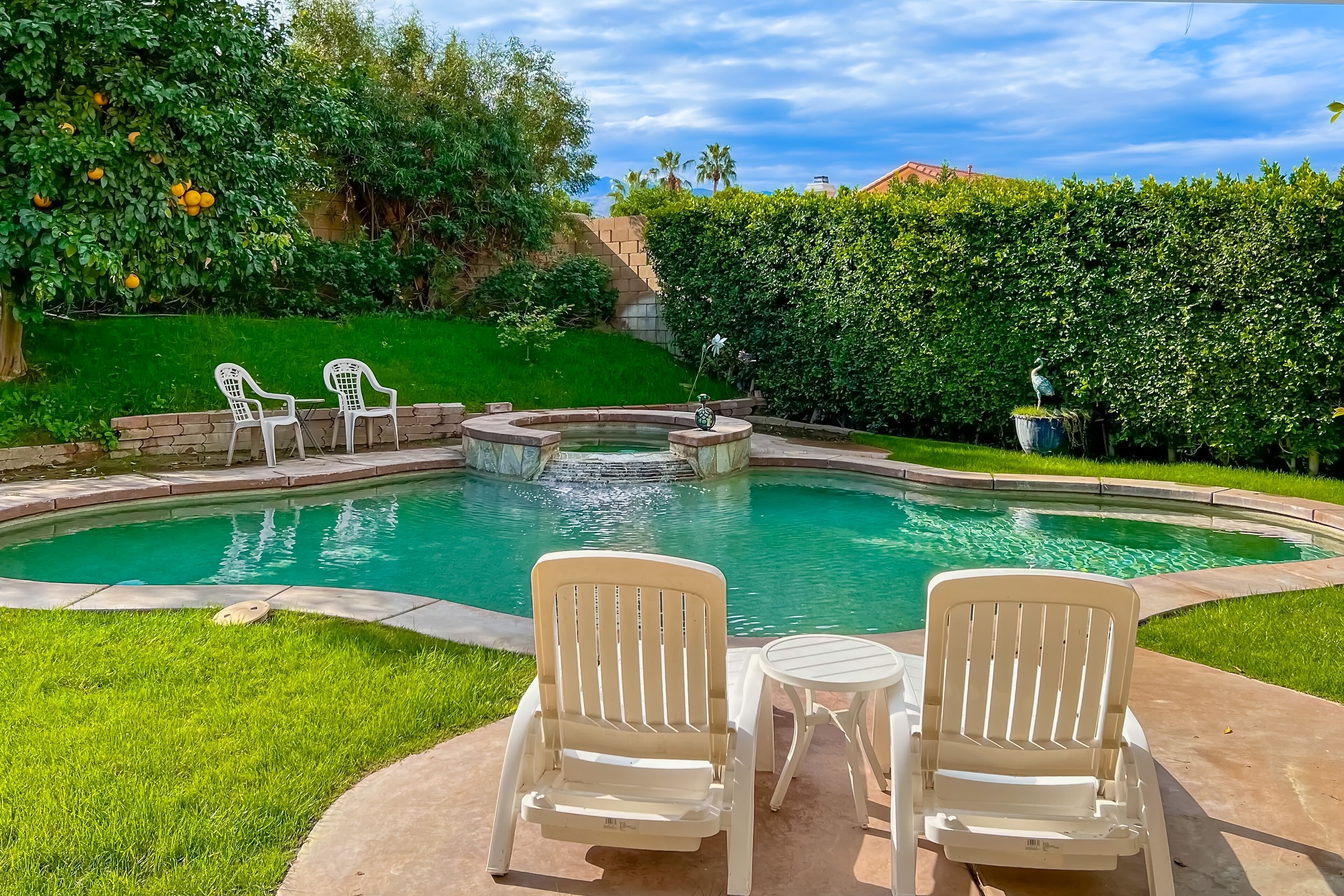 A swimming pool with two white chairs and a small table, surrounded by a well-maintained lawn and hedges.