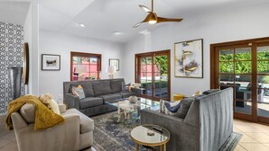 Un salon spacieux avec un canapé gris, un canapé beige et une table basse en verre. Il y a deux fenêtres avec des cadres en bois, un ventilateur de plafond et un grand tableau accroché au mur.