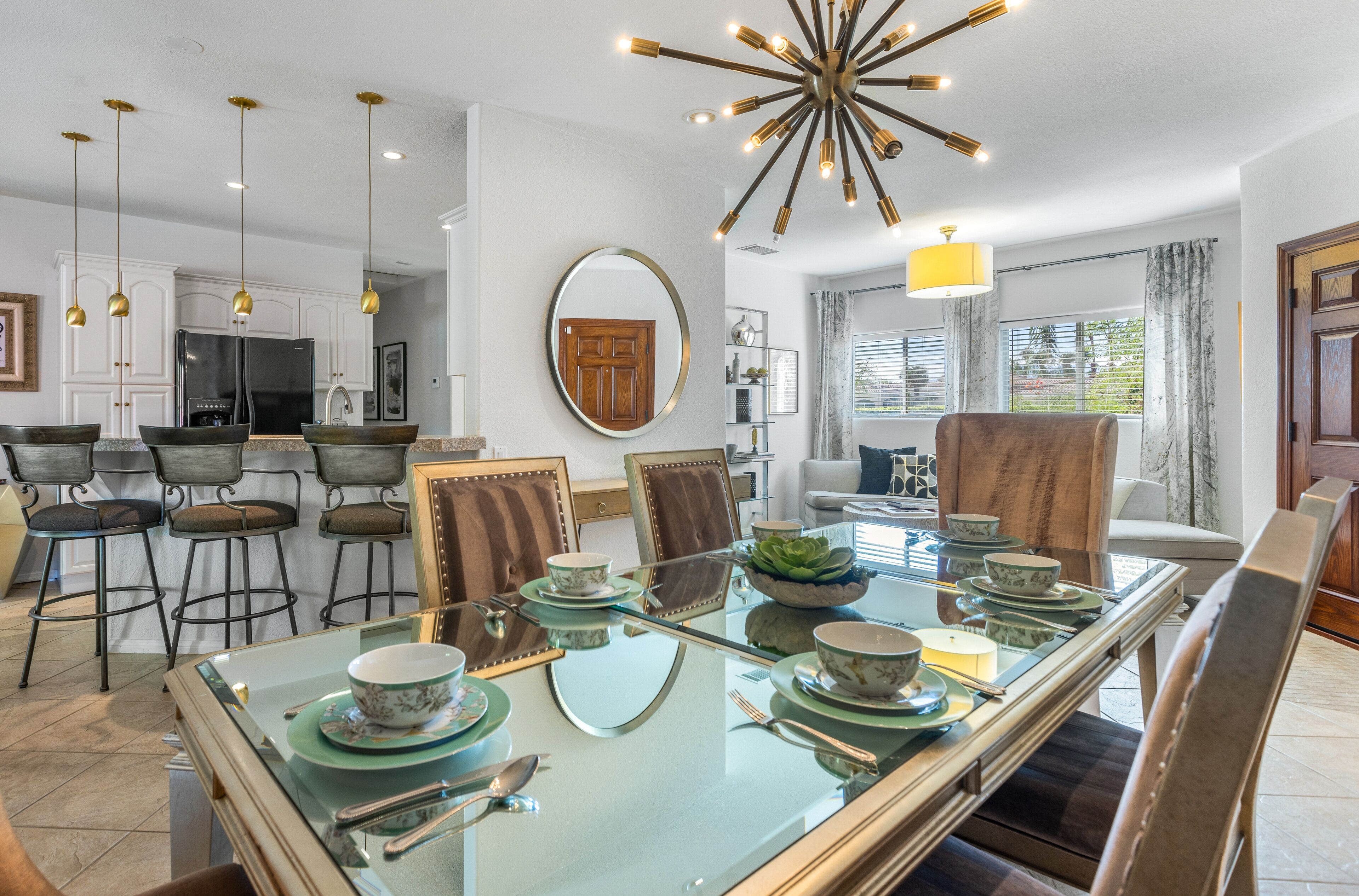 A modern dining area with a glass table set for six, surrounded by wooden chairs. A kitchen with white cabinets and a refrigerator is visible in the background.