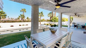 An outdoor patio with a table and chairs, a ceiling fan, and a view of palm trees and a swimming pool. - Mountain-View Haven with Pool, Peloton, Luxury Oasis near El Paseo (Palm Desert)