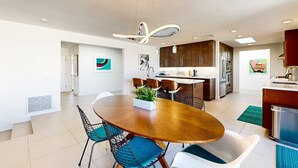 A modern kitchen with dark wooden cabinets, a central island, and a dining area with a round wooden table and blue chairs. - Mountain-View Haven with Pool, Peloton, Luxury Oasis near El Paseo (Palm Desert)