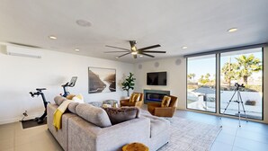A modern living room with a grey sofa, a ceiling fan, a mounted TV, and a large window offering a view of palm trees. - Mountain-View Haven with Pool, Peloton, Luxury Oasis near El Paseo (Palm Desert)
