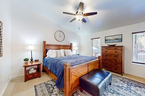 Une chambre à coucher avec un lit en bois, une table de chevet, une commode, un ventilateur de plafond, une horloge et une fenêtre avec des stores.