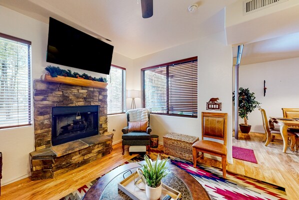 A living room with a stone fireplace, a flat-screen TV mounted above it, a comfortable armchair, a wooden bench, and a dining area with a table and chairs.