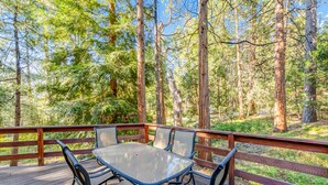 Une terrasse en bois, dotée d’une table et de chaises à plateau en verre, entourée d’une balustrade en bois, et offrant une vue sur une zone boisée.