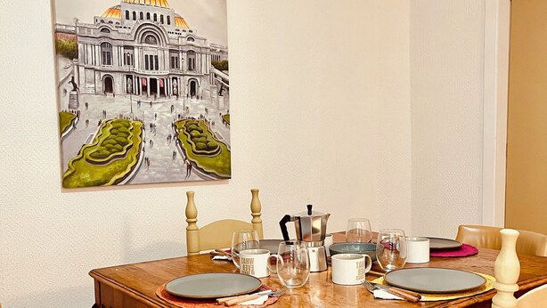 Ein Essbereich mit einem Holztisch, gedeckt für vier Personen, umgeben von Stühlen. Ein Gemälde eines Gebäudes mit einer Kuppel hängt an der Wand.