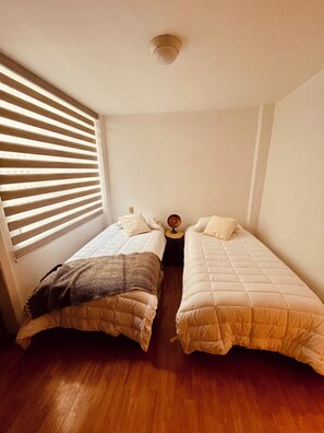 Ein kleines, gemütliches Schlafzimmer mit zwei Einzelbetten, einem Nachttisch mit Lampe und einem Fenster mit Jalousien.