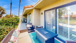 A patio with a sofa, a table, and a chair. There are palm trees and a hedge in the background. - Palm Desert Resorter | New Furnishings | 20 Pools, Championship Golf (Palm Desert)