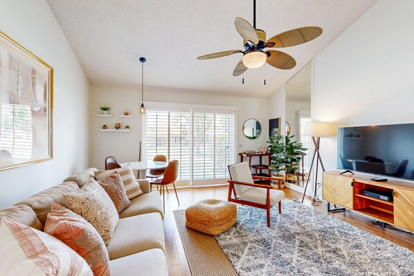 A modern living room with a sofa, a television, a ceiling fan, and a dining area with chairs and a table. - Palm Desert Resorter | New Furnishings | 20 Pools, Championship Golf (Palm Desert)