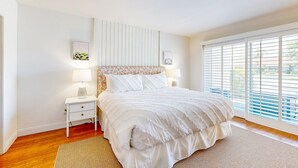 A bedroom with a large bed, a nightstand, a lamp, a wall-mounted shelf, and a window with blinds. - Palm Desert Resorter | New Furnishings | 20 Pools, Championship Golf (Palm Desert)
