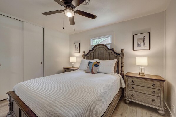Un dormitorio con cama, mesitas de noche, ventilador de techo y obras de arte colgadas en la pared.