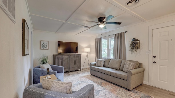 Ein Wohnzimmer mit Deckenventilator, einer Couch, einem Sessel, einer Lampe und einem großen Fenster mit Vorhängen.