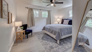 Ein Schlafzimmer mit Bett, Deckenventilator, Fenster mit Vorhängen, Nachttischlampe und einem kleinen Tisch mit Pflanze.