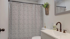 Ein Badezimmer mit gemustertem Duschvorhang, Toilette, Waschbecken mit Spiegel und einer kleinen Pflanze an der Wand.